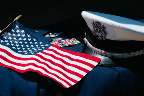 A white hat with an eagle emblem is placed on a dark surface next to an American flag and a collection of medals.