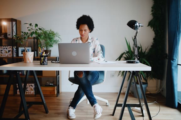 A person is sitting at a desk with a laptop, surrounded by plants and a lamp.