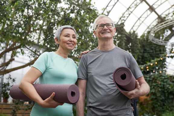 A man and a woman are holding yoga mats and standing in a greenhouse.