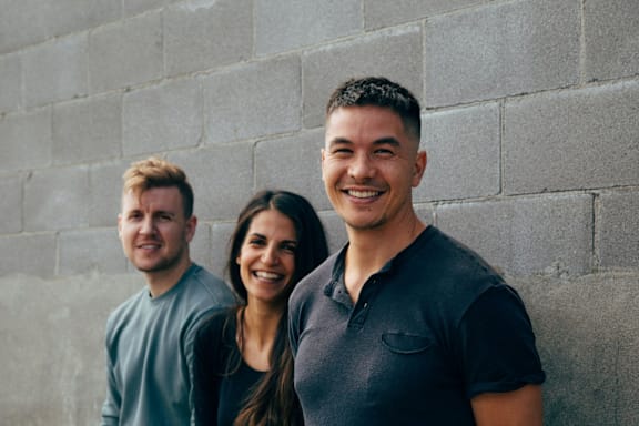 Three people smile at the camera against a grey wall.