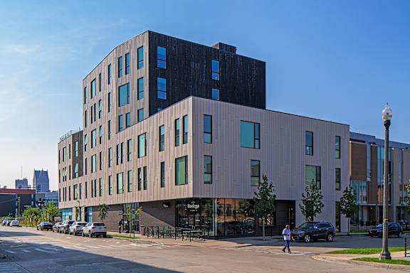 A modern building with a black top floor and tan bottom floors with a person walking in front of it.
