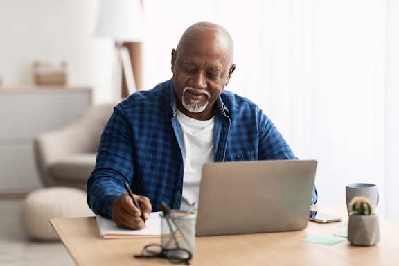 A man in a blue shirt is writing in a notebook while using a laptop.