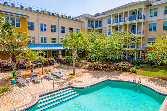 A swimming pool is surrounded by a patio with chairs and a building with balconies.