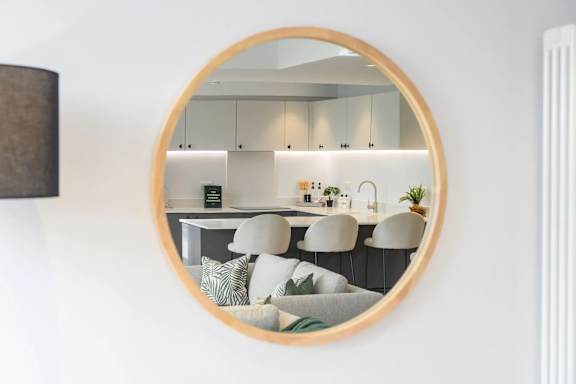A round mirror reflects a modern kitchen with white cabinets and a dining area.