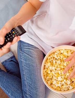 A person is sitting on a couch holding a remote control and a bowl of popcorn.
