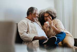 Two people sitting on a couch, one holding a cup and the other holding a book.