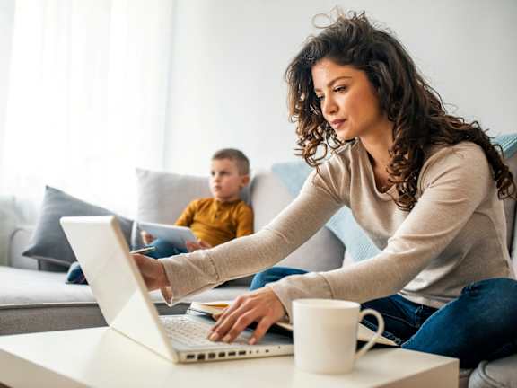 Woman Sitting on Sofa Working on Laptop while Son Sits Next to Her