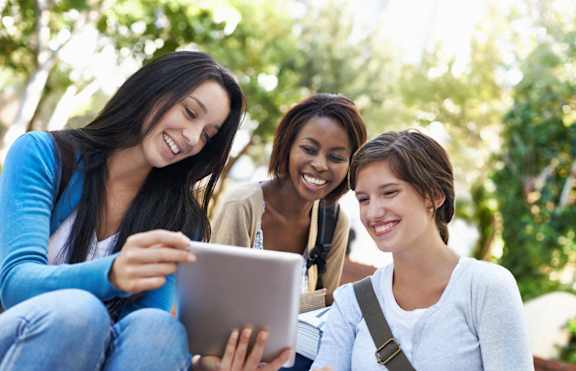 Three women are sitting outdoors and looking at a tablet.