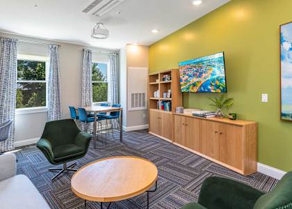The Residences at Crosspoint cyber lounge in Lowell, MA includes a green accent wall with a mounted flat-screen TV, a wooden storage unit with open shelving, a dark green swivel chair, a round wood coffee table, and a high-top table with blue chairs by two curtained windows overlooking trees.