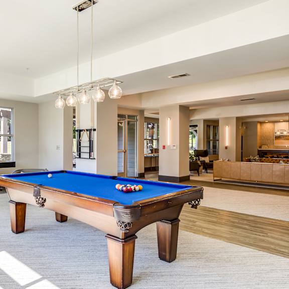 A pool table with a blue cloth and white balls is in the center of a room with a high ceiling and a chandelier.