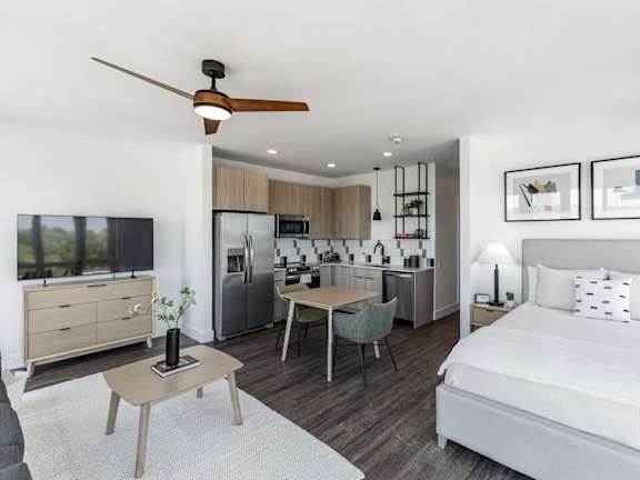 A modern bedroom with a bed, a table, a ceiling fan, and a kitchen area in the background at Anthem House Apartment, Huntsville, AL, 35806