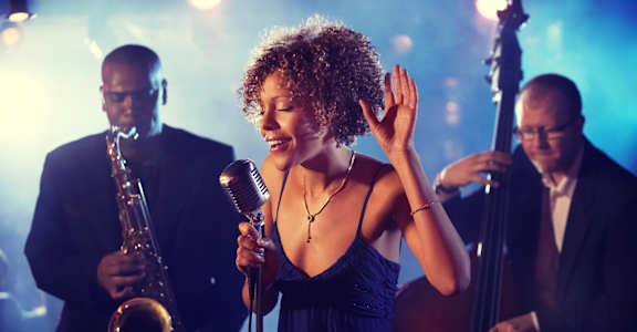 A woman in a blue dress is singing into a microphone with a saxophonist and a cellist in the background.