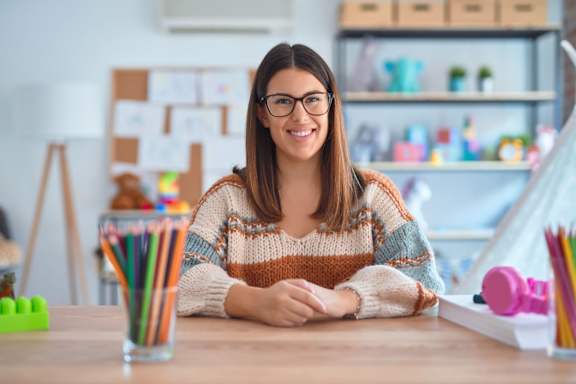 A woman with glasses is sitting at a desk with a glass of colorful pencils in front of her.