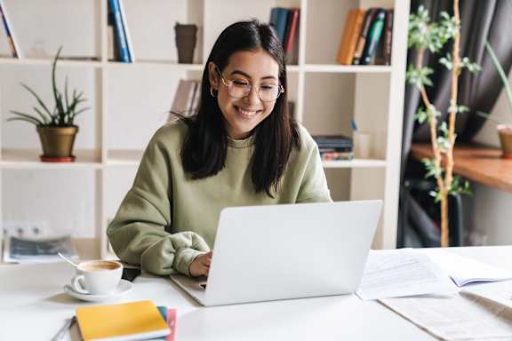 A woman in a green sweater is working on her laptop at a desk with a cup of coffee and some books.