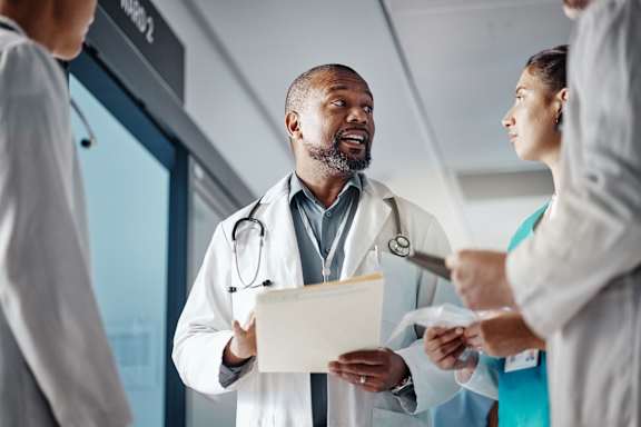 A group of medical professionals are gathered around a doctor who is holding a clipboard.