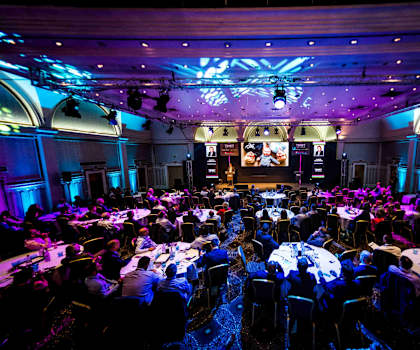 A large conference room with people seated at tables facing a stage.