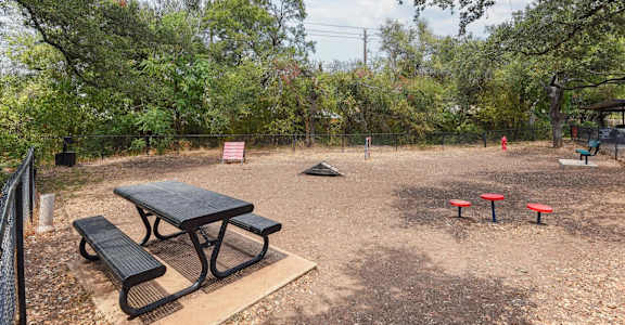 A picnic table and two red stools are in a fenced-in area with trees in the background.