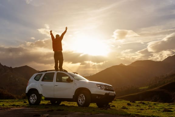 Man Standing on Car with Arms Up Admiring Sunset