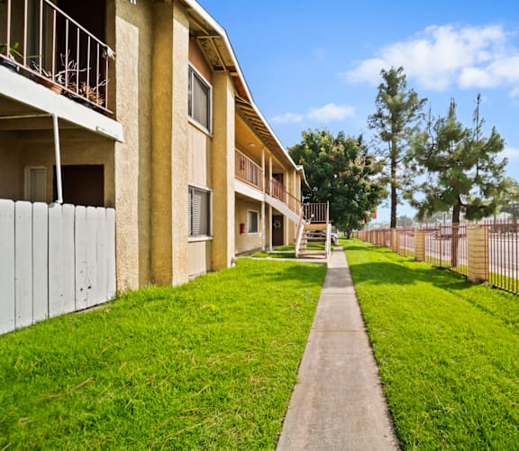 A long concrete walkway leads to a building with a metal fence on one side and a grassy area on the other.
