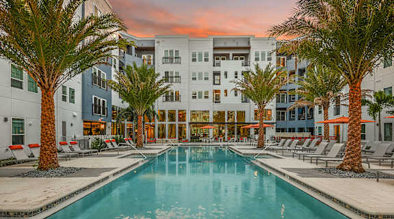 A swimming pool surrounded by palm trees and lounge chairs in front of a multi-story building.
