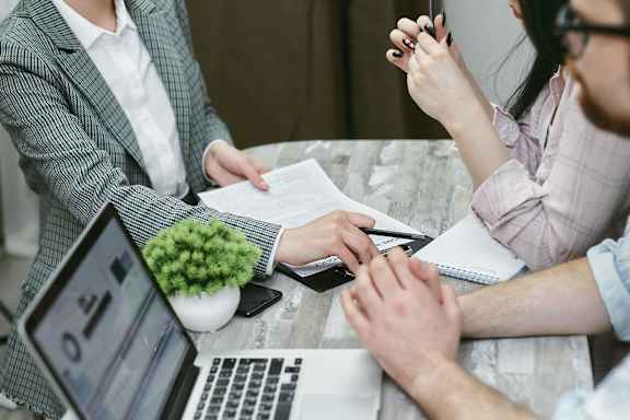 Two people sitting at a table with laptops and papers, one person is writing on a notepad.