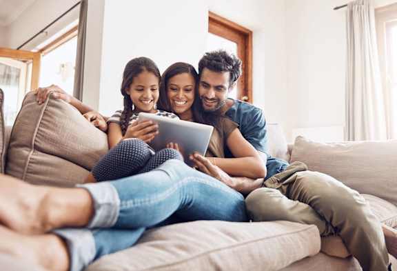 A family of three is sitting on a couch, looking at a tablet together.