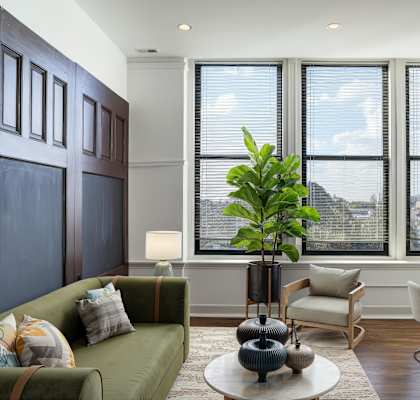 A living room with a green couch, a table with a vase and a potted plant, and a view of the city through the windows.