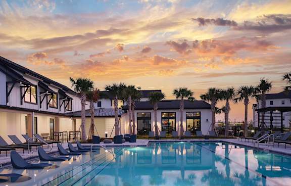 A pool surrounded by lounge chairs and palm trees under a sunset sky.