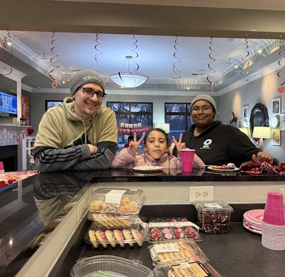Three people are standing behind a counter with various food items on it.