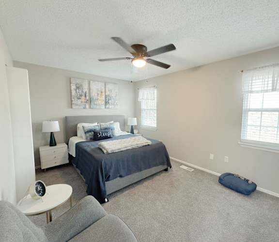 Reserve at Hillcrest model unit staged bedroom with blue blankets, gray chair, two windows with curtains and a dog bed