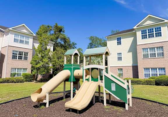 A playground with a green slide and a white railing in front of apartment buildings.