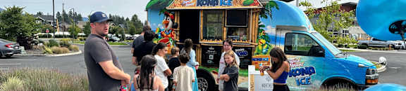 A food truck with a colorful design is parked on a paved area with people standing in line to order.