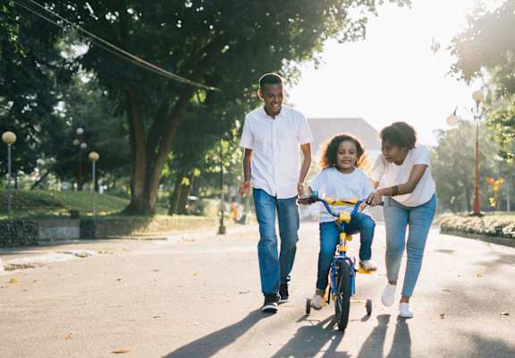 A female child rides a bike while a female adult holds the handlebars and a male adult follows behind