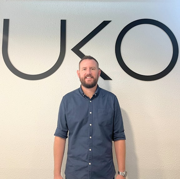 A man with a beard and short hair is standing in front of a wall with the letters U, K, and O.