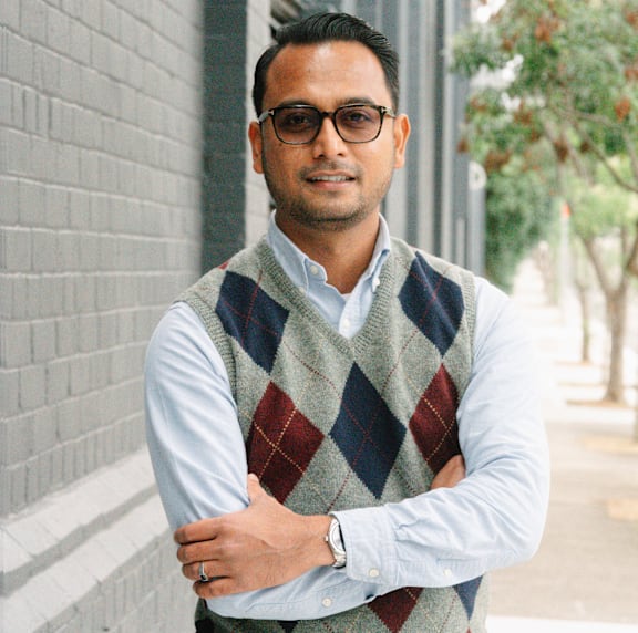 A man wearing glasses and a sweater vest stands with his arms crossed.