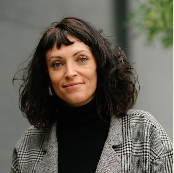 A woman with short dark hair and a black turtleneck is wearing a grey and white checkered blazer.