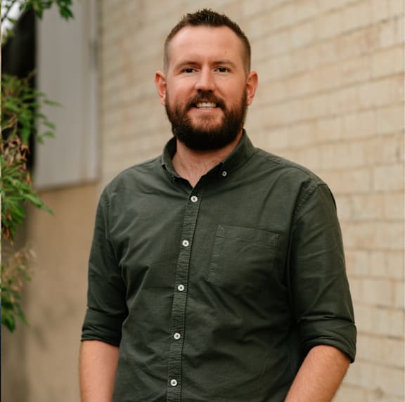 A man with a beard is standing in front of a brick wall.
