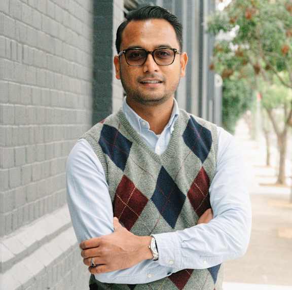 A man wearing glasses and a checkered vest stands with his arms crossed.