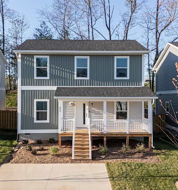 A modern two-story home with a green exterior, white trim, a covered front porch, and a neatly landscaped yard in a suburban neighborhood.