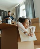 A young girl is sitting in a cardboard box in a room with a man and another person in the background.