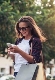 Woman Smiling while Looking at Phone and Carrying Shopping Bags