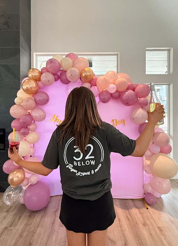 A woman in a black t-shirt with the text "32 Below" is standing in front of a balloon arch.