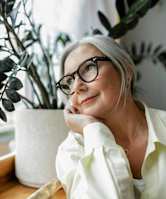 A woman with glasses and a white shirt is sitting in front of a plant.