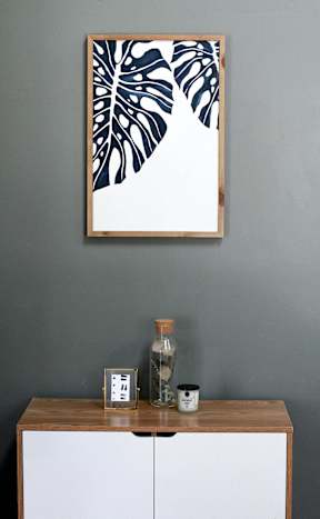 A white cabinet with a wooden top and a framed leaf pattern picture on the wall above it.