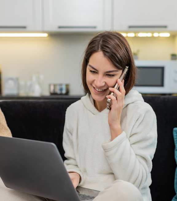 A woman is sitting on a couch with a laptop and talking on the phone.
