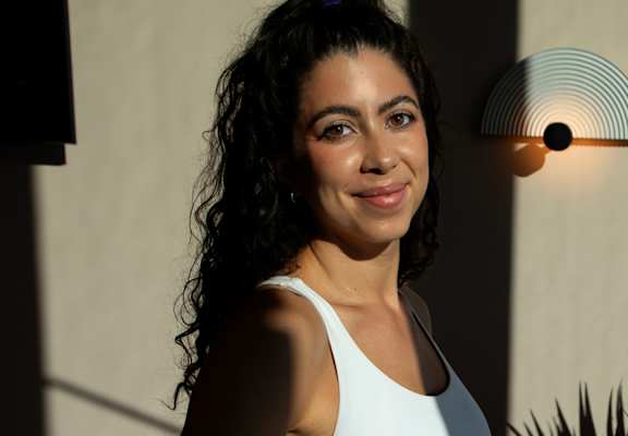 A woman with dark hair and a white tank top stands in front of a wall with a light fixture.