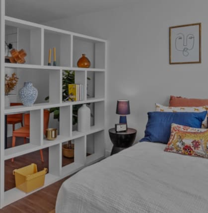 A bedroom with a bed, a table, and a shelf with various items on it.