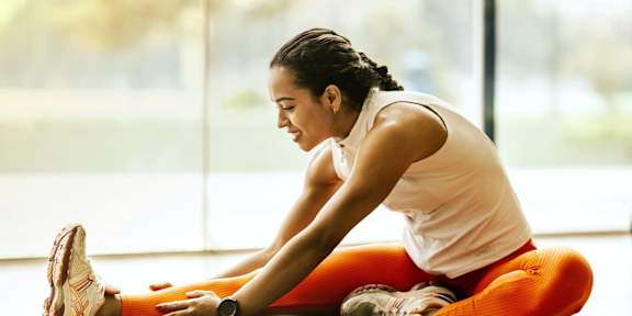 A woman in a white top and orange leggings stretches her leg while sitting on the floor.