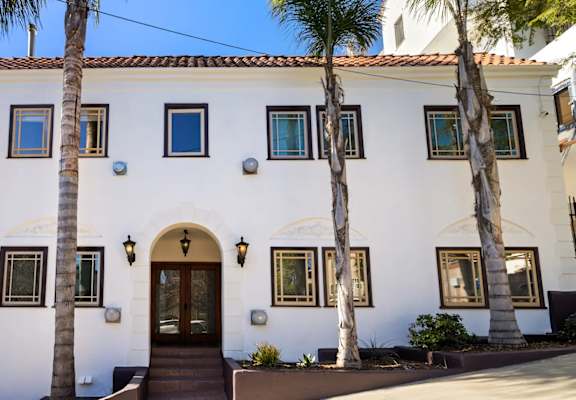 A white house with a brown door and windows surrounded by palm trees.