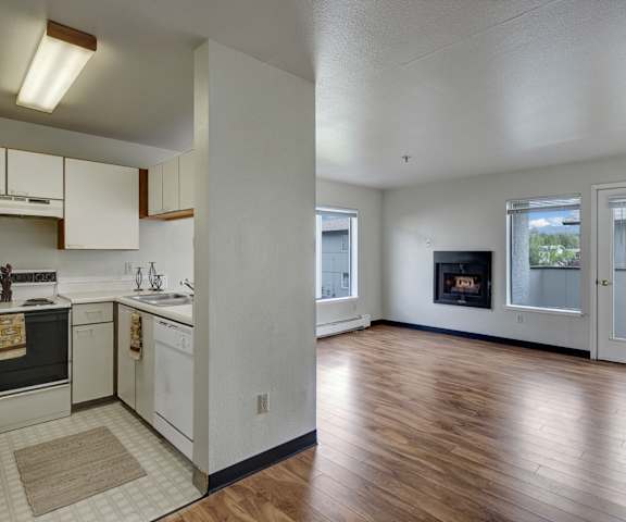 A view of the large empty living room with windows, a fireplace, and a door the balcony and the kitchen with white appliances and white wooden large cabinets at Silver Ridge Apartment Homes. 